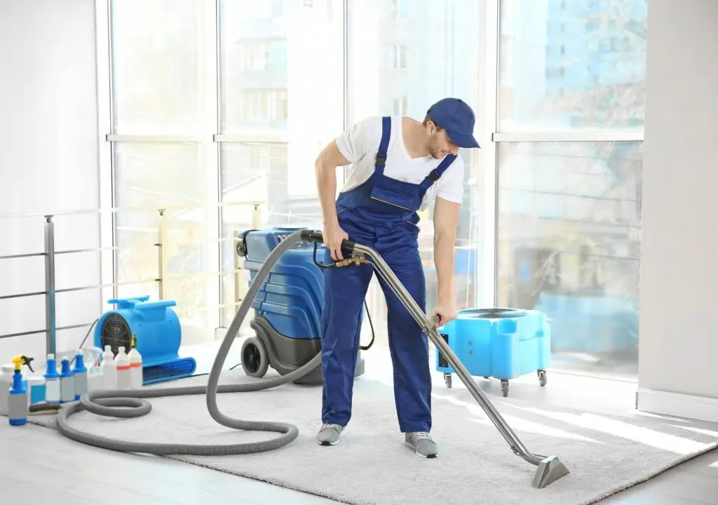 A man in blue overalls and a cap expertly uses the best vacuum cleaner to tidy a beige carpet in a bright room with large windows. Nearby, cleaning supplies and equipment are neatly arranged for optimal carpet cleaning results.