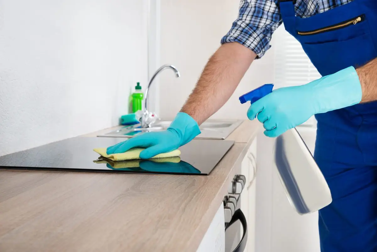 A person wearing blue gloves and overalls diligently cleans a kitchen countertop with a spray bottle and cloth. In the background, a sink sits beside a green dish soap bottle and crumpled foil, hinting at the best tile cleaning practices in action.