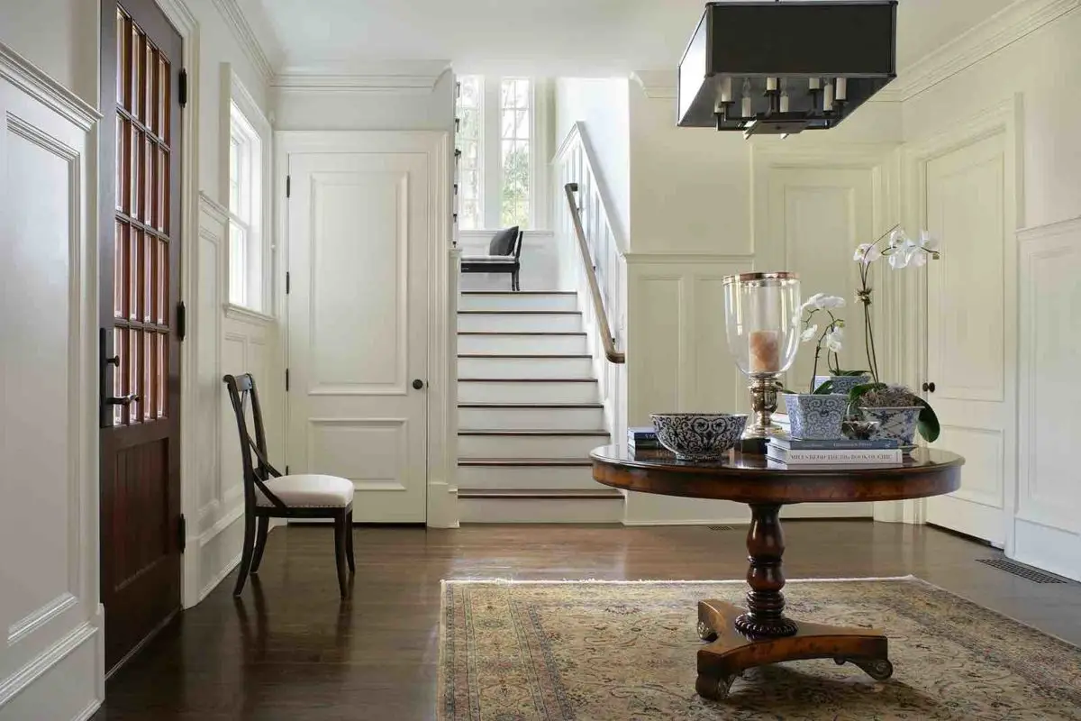 A bright foyer with a wooden staircase features an ornate round table adorned with decorative items and a chair with a cushioned seat. White paneled walls and a large square light fixture accentuate the space, while a top-rated carpet cleaning service ensures the patterned rug remains pristine.