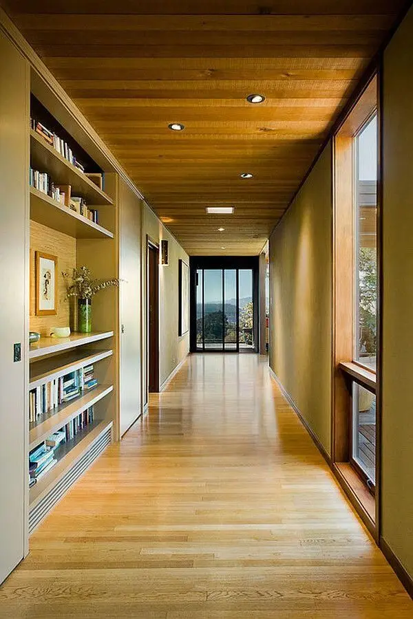 A modern hallway with top-rated wooden floors and ceiling features built-in shelves filled with books and decor on the left. Large windows on the right let in natural light, leading to a glass door at the end with a view outside.