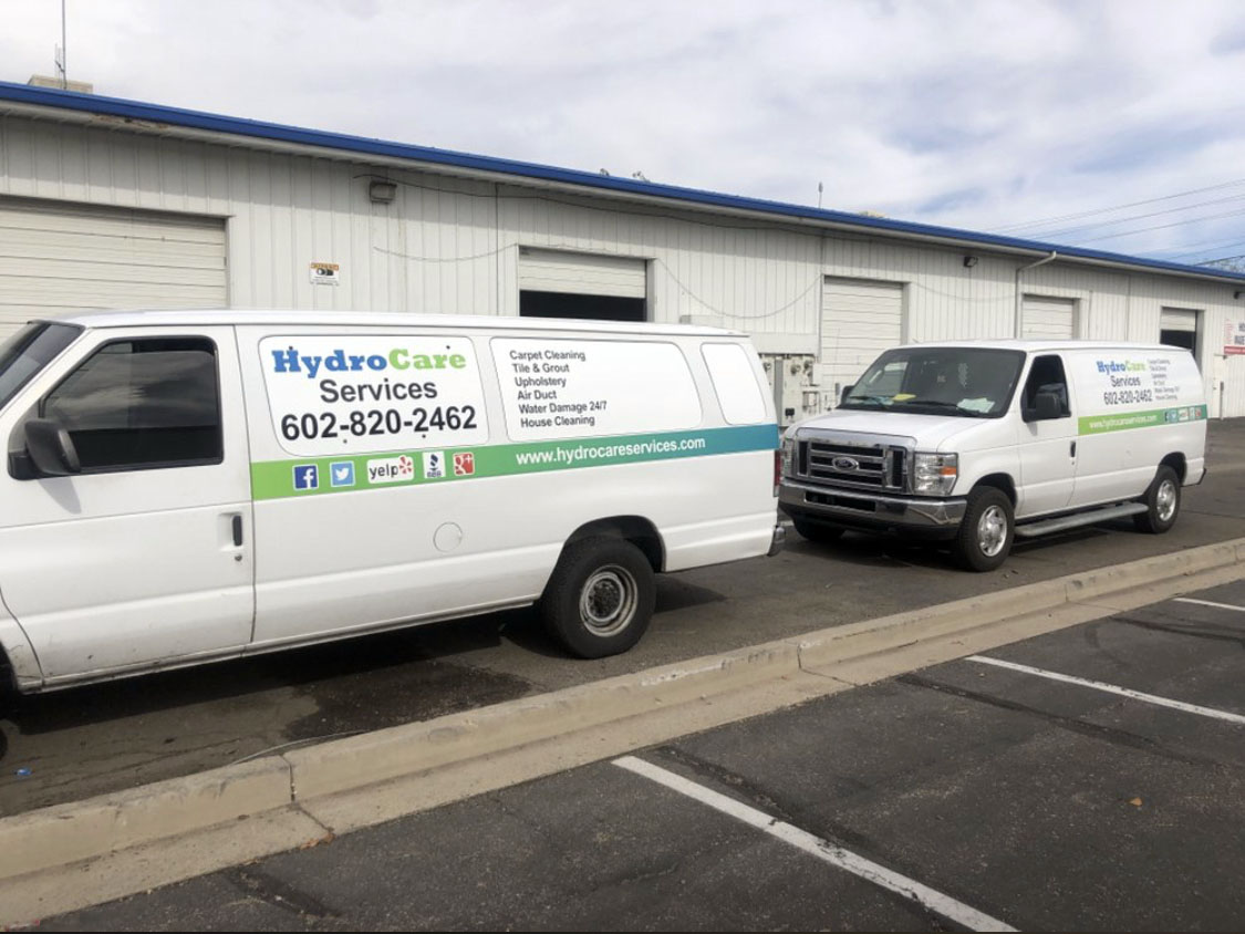 Two white commercial vans for HydroCare Services parked side by side, with company branding and contact information, offering carpet cleaning and other services in Phoenix