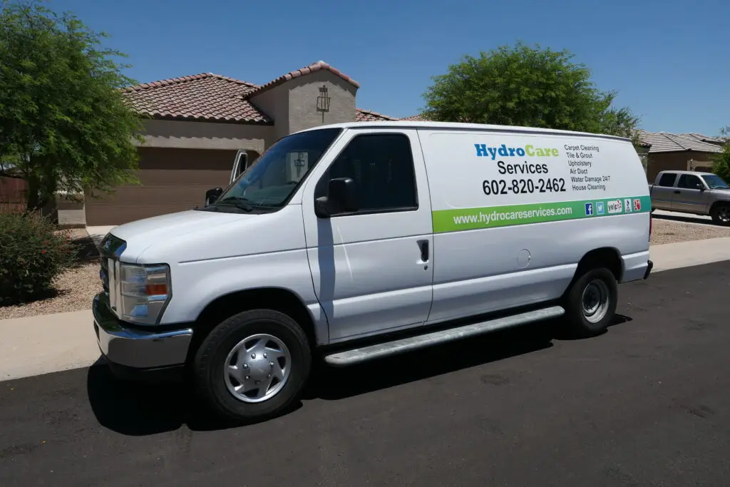A white service van parked on a residential street. The van features HydroCare Services branding, contact number, and a list of services including carpet cleaning and tile cleaning. Background includes desert-style homes and trees.