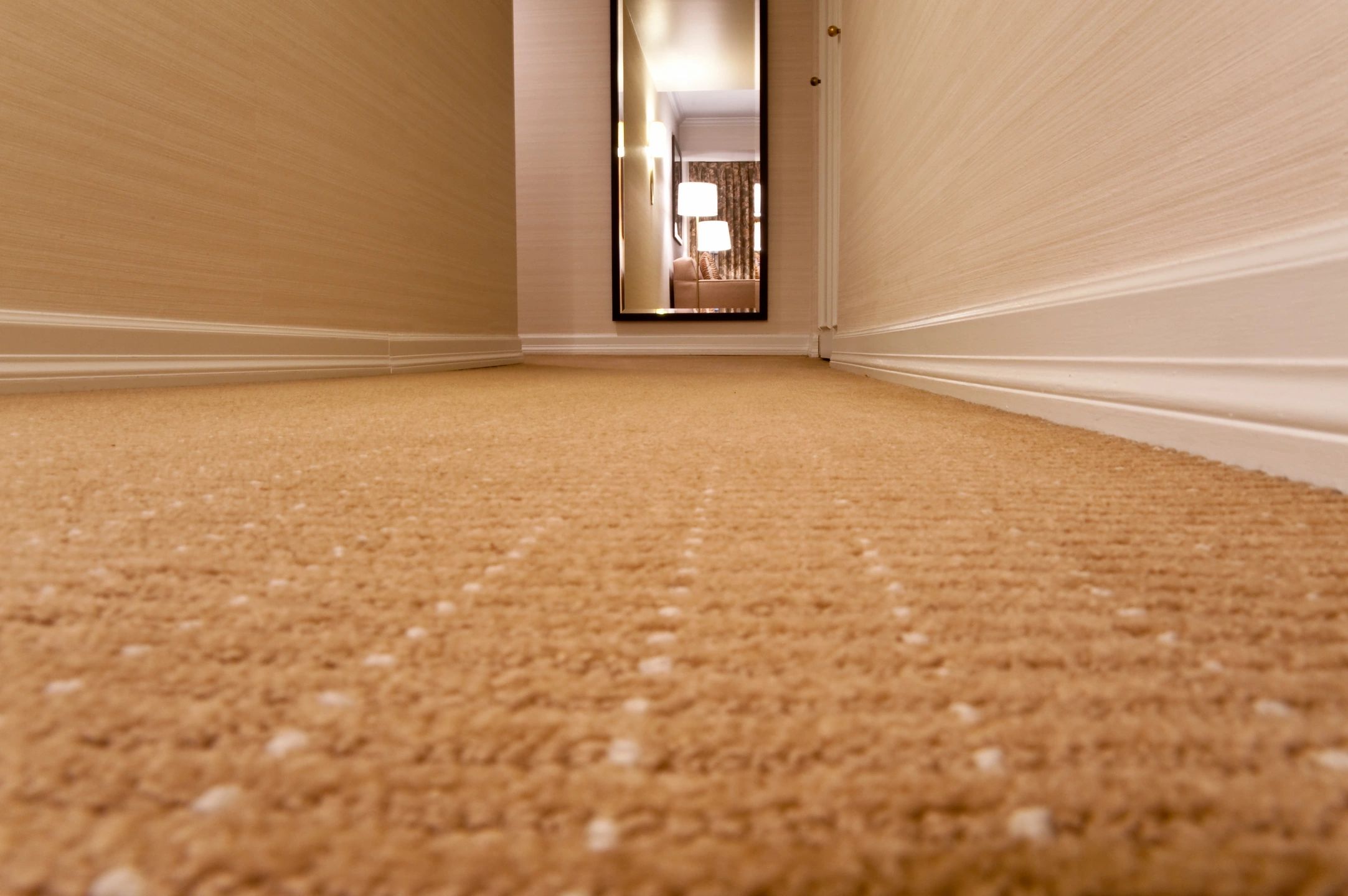 Low-angle view of a long, beige-carpeted hallway with cream-colored walls, leading to an open door revealing a lit room with a bed and lamp in the distance.