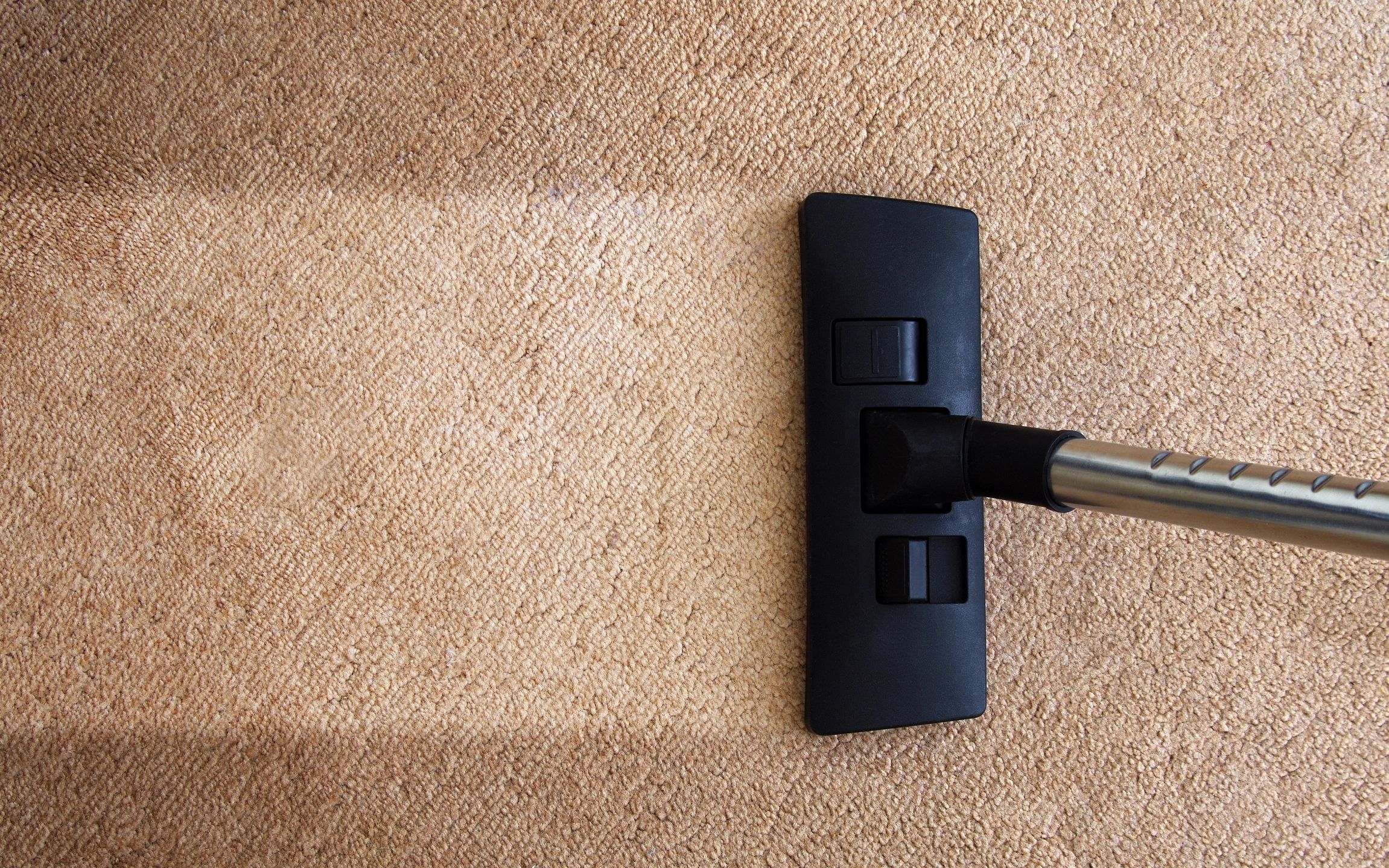 A vacuum cleaner head is cleaning a beige carpet, leaving a visible clean path behind it. The difference between the cleaned and uncleaned sections of the carpet is noticeable.