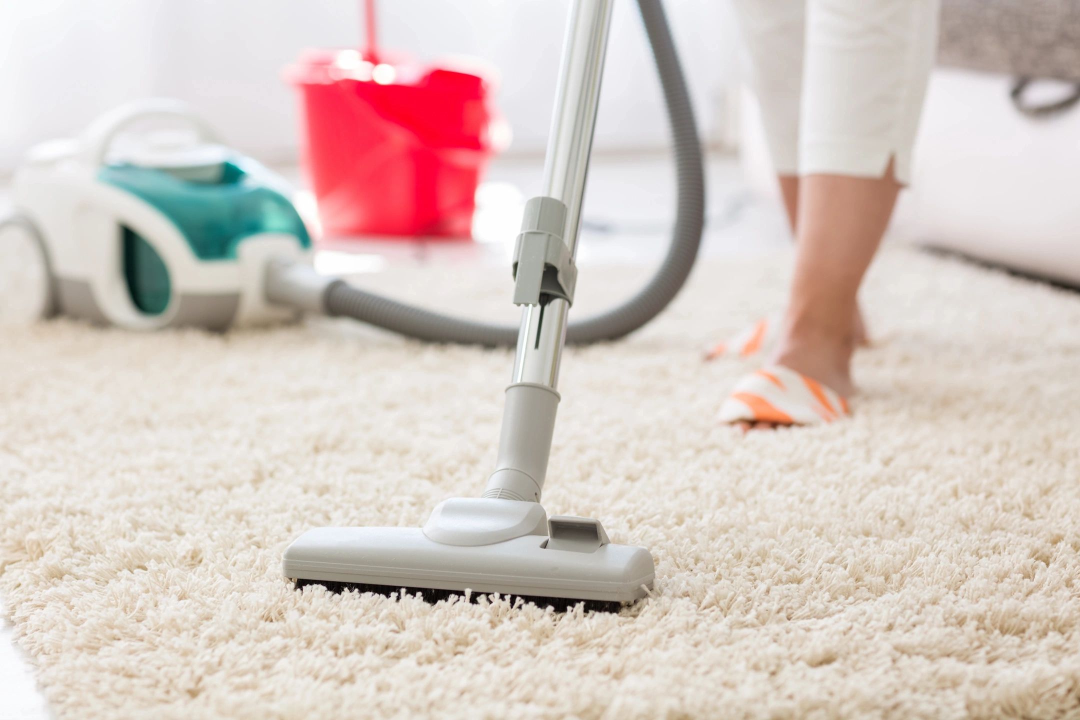 A person vacuuming a light-colored carpet in a bright room, with a canister vacuum cleaner and a red bucket visible in the background. Only the lower legs and feet of the person are shown.