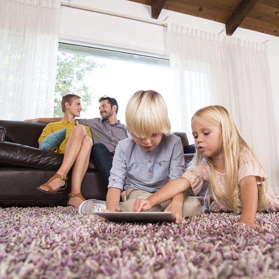 Brother and sister using tablet on carpet in living room with parents in background