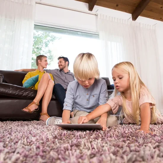 A boy and a girl sit on a carpet, engaged with a tablet. In the background, a man and woman relax on the couch, smiling and talking. The room is brightened by natural light through large windows—truly the best setting for family time.